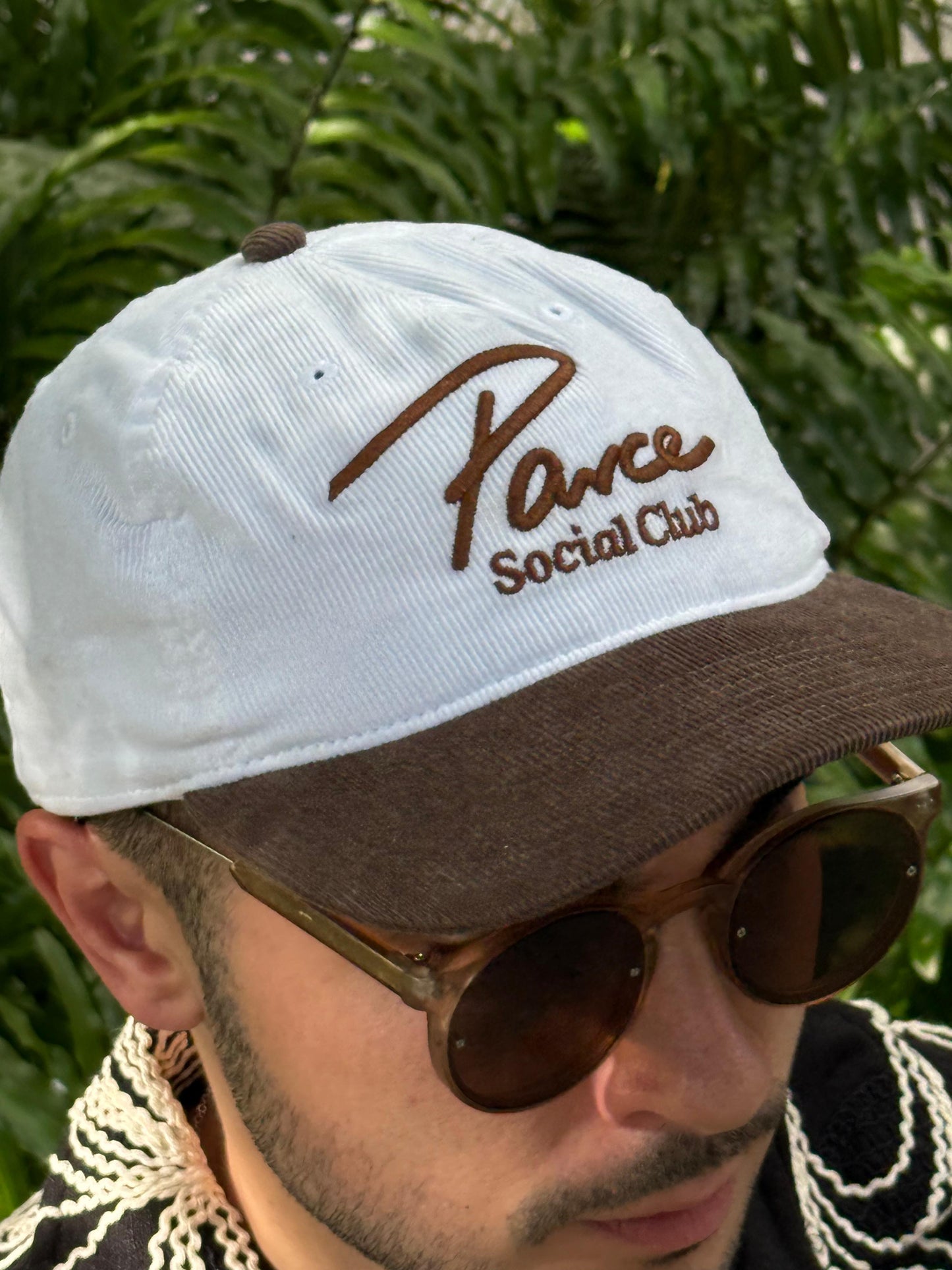 Man wearing Parce Social Club corduroy hat with white crown, brown brim, and brown embroidered logo, standing in front of green foliage.