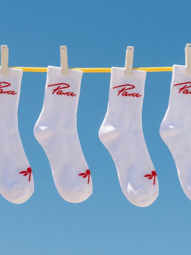 Four white Parce cotton crew socks featuring the red script logo and signature palm tree icon, hanging on a clothesline against a clear blue sky.